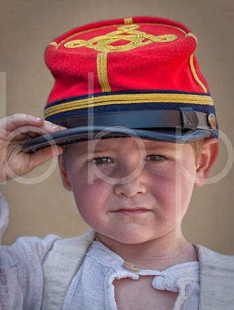 A little boy wearing a red hat and a dirty white shirt salutes passing civil war reenactment troops as they march off to war.