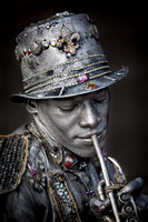A New Orleans French Quarter Mime Wearing A Jewel Encrusted Top Hat And Adorned in Silver Paint Who Feigns Playing A Keyless Trumpet is shown in this environmental portrait photograph by Brian Buckner