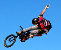 A young and colorfully dressed bicycle riding acrobat jumps high in the air while performing stunts in a half-pipe course under a blue cloudless sky.