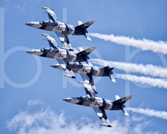 The Black Diamond Jet Team with white smoke billowing from their engines fly in formation at 600 miles per hour above the cheering crowd and under a blue sky with white clouds at an airshow at Barksda