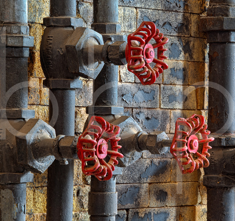 Three red valve wheels and the grey painted pipes they are attached to are contrasted by the chipped paint of a wall in this commercial architectural photograph by Brian Buckner Photography, Shrevepor