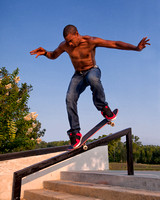 A young, bare-chested or shirtless black man wearing jeans and red tennis shoes performs a skateboard trick by riding on a stair rail using only the front two wheels of his skateboard.