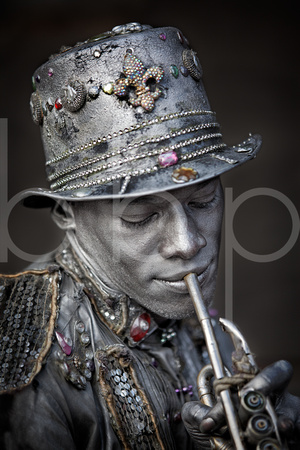 A New Orleans French Quarter Mime Wearing A Jewel Encrusted Top Hat And Adorned in Silver Paint Who Feigns Playing A Keyless Trumpet is shown in this environmental portrait photograph by Brian Buckner