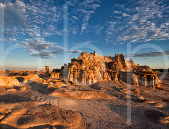 A beautiful sandstone formation in the bisti/de-na-zin wilderness area that looks like a castle made of clay and rock is illuminated by the rays of the dawning sun under a blue sky with striated cloud