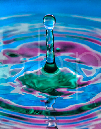 A single water drop strikes a container with green water in it causing a large column of water to rise reminiscent of an atomic blast.  Colorful blue and red gels covering strobe lights create a color