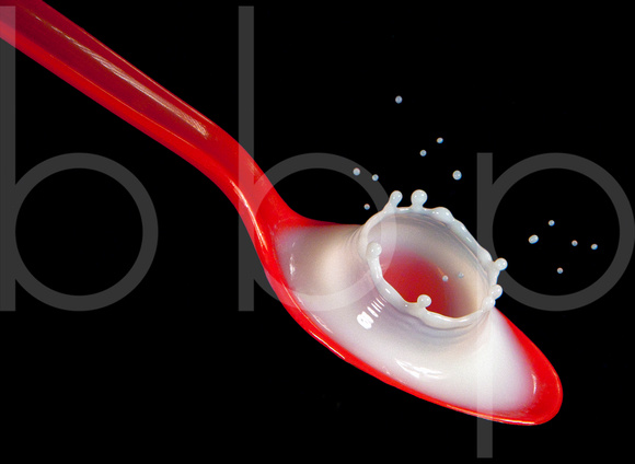 A drop of milk impacts a red spoon filled with more milk creating a corona or crown like splash in this commercial product photograph by Brian Buckner Photography, Studio "B", Shreveport, Louisiana.