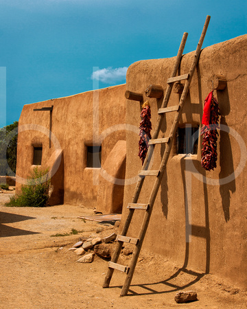 Red chile pepper ristras hang by a window decorating an adobe dwelling as a leaning wooden ladder casts shadows on the wall under a turquoise sky.