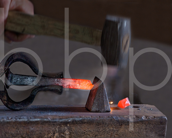 A blacksmith's hammer strikes and cuts through hot glowing iron on the anvil in a forge in this commercial environmental industrial photograph by Brian Buckner Photography, Shreveport, Louisiana.